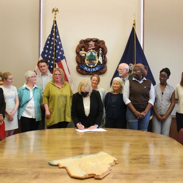 Group of people gathered around a large wood table. The person in the center signs a piece of legislation